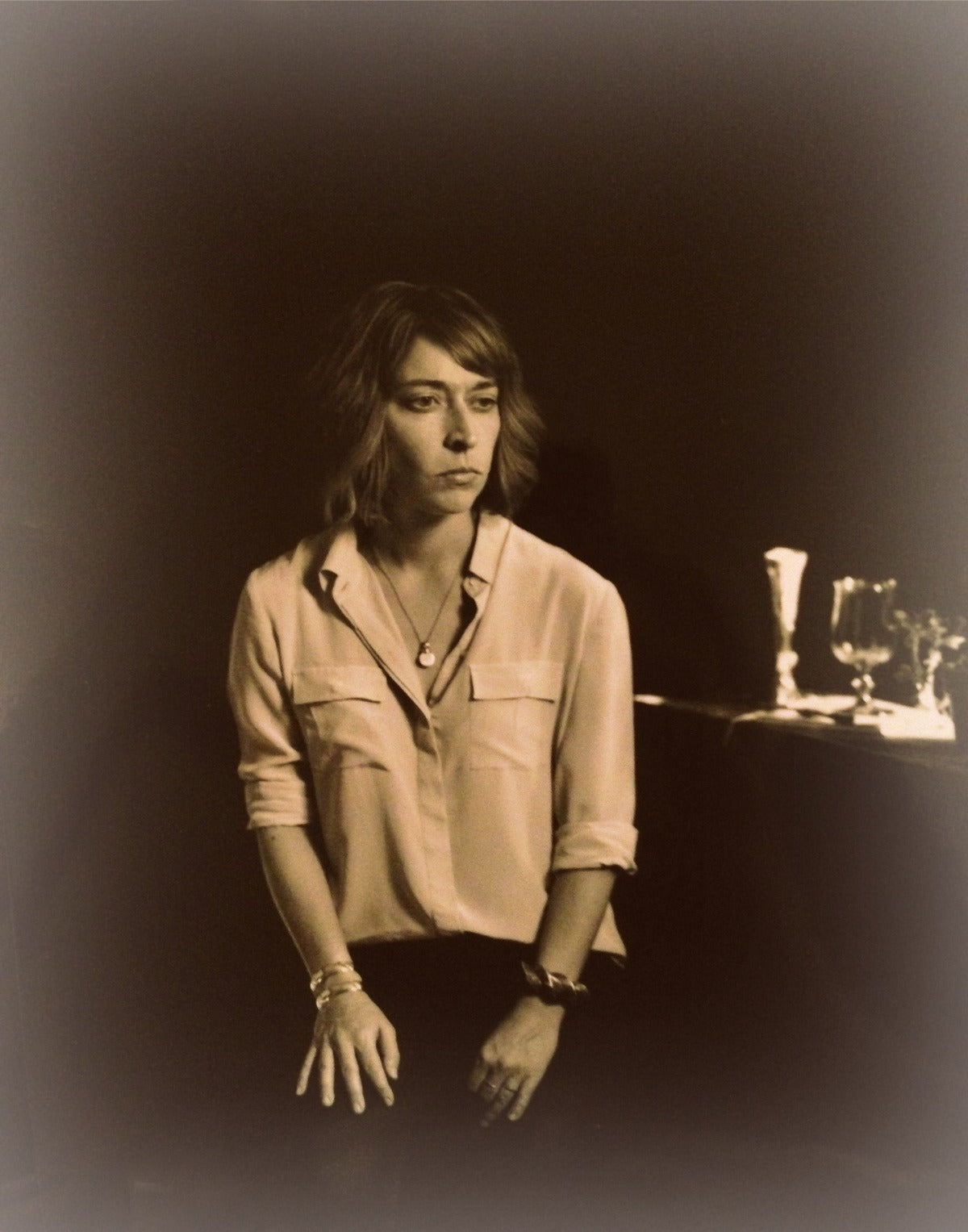 Sepia-toned portrait of a woman sitting indoors near glassware on a table, artistic style.