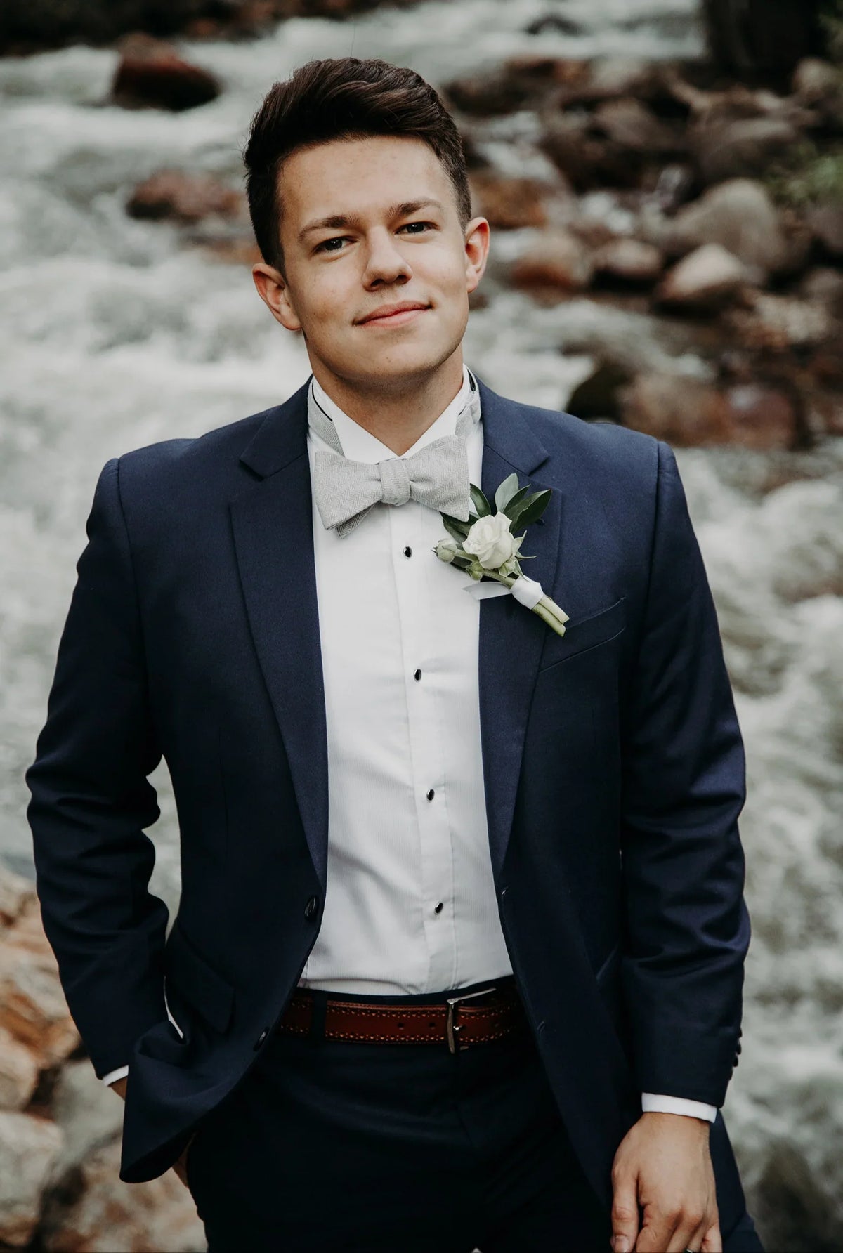 Man in formal suit with boutonniere and bow tie standing by a rocky river outdoors