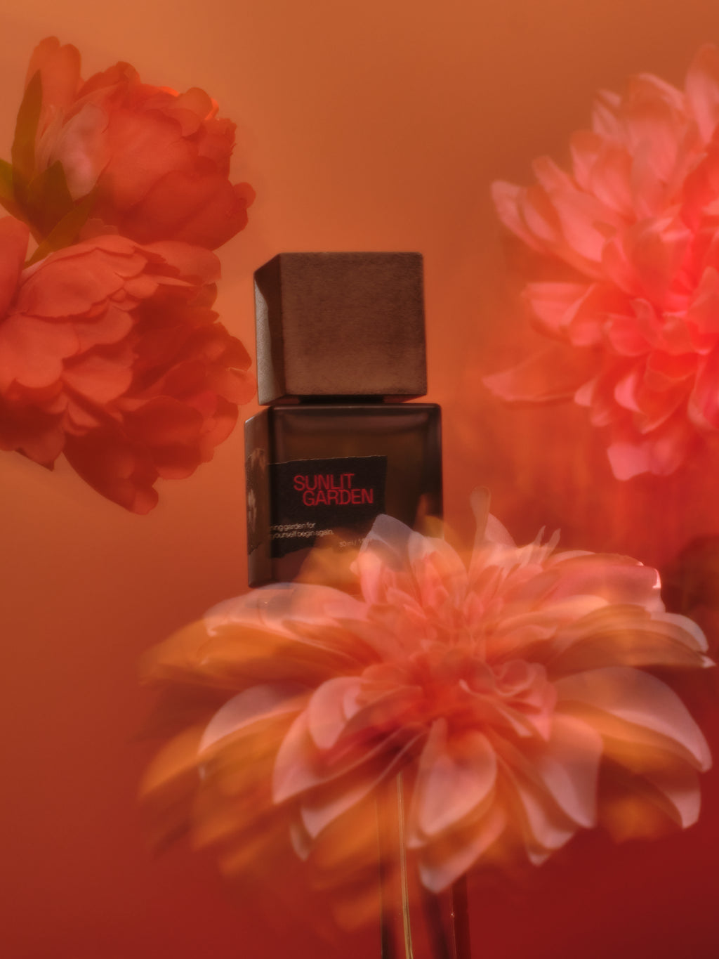 A bottle of Sunlit Garden perfume centered against a warm orange background with large blurred flowers in the foreground and background.